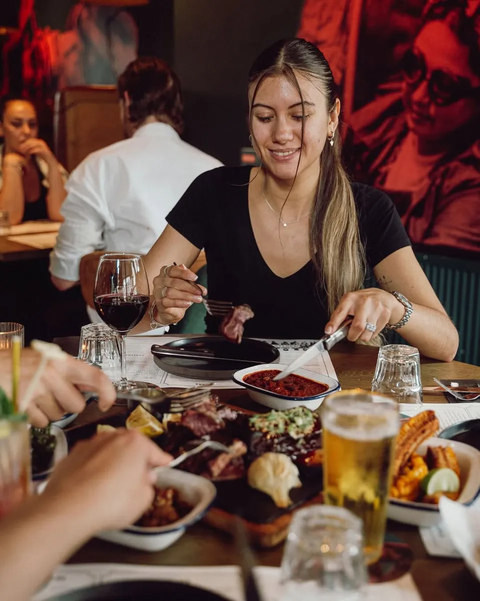 A guest smiling over a forkful of steak beside a loaded sharing board of cuts and sides
